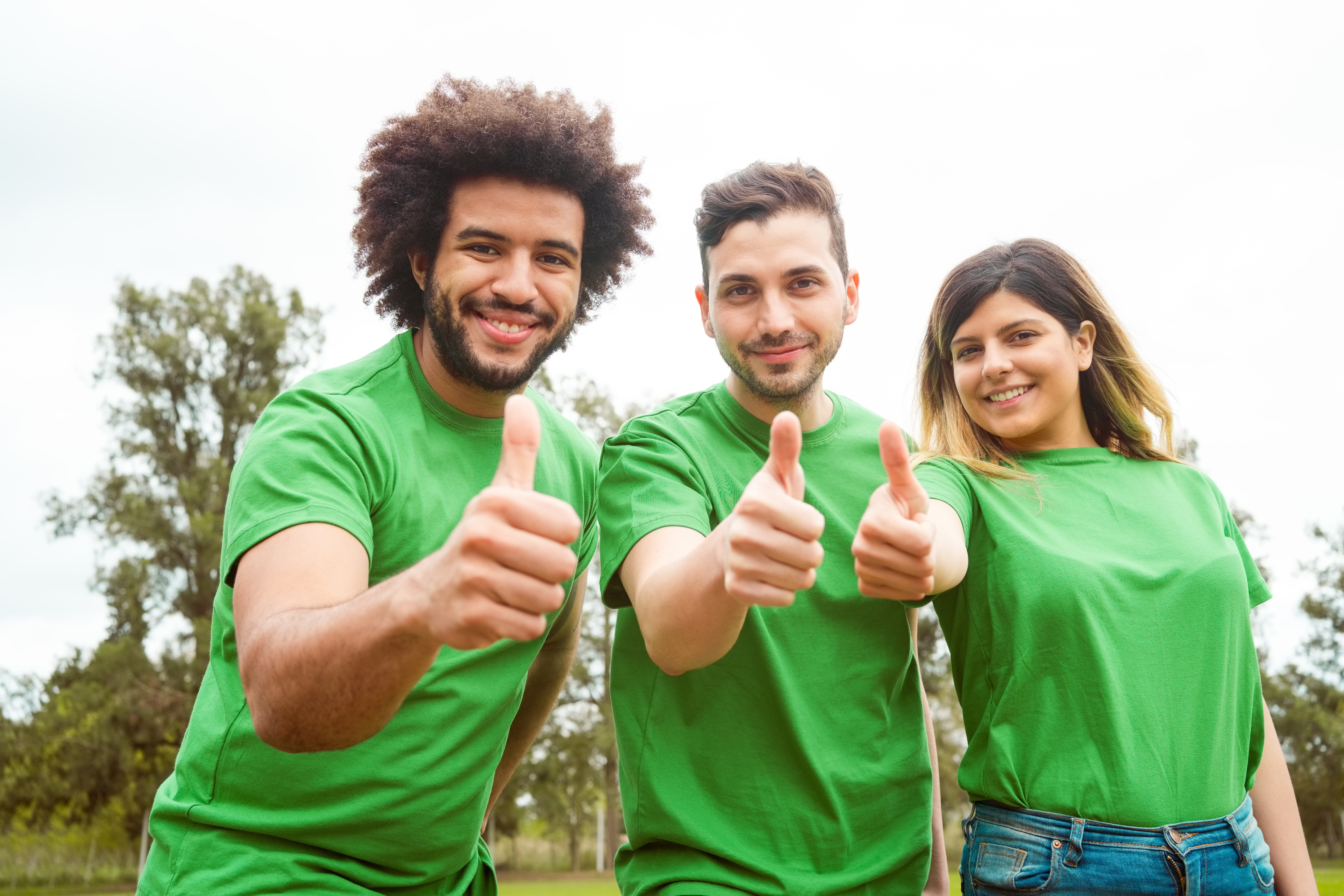 Portrait of smiling male and female volunteers showing thumbs up. Confident environmentalists are in public park. They are in green t-shirts.