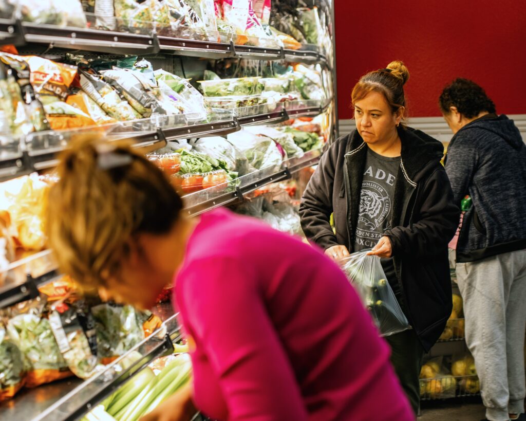 A woman in a grocery store collects zucchini in a bag from a wall of fresh veggies and greens